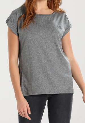 Kvinde iført en afslappet kortærmet grå t-shirt og sorte bukser, stående mod en ensfarvet lys baggrund.