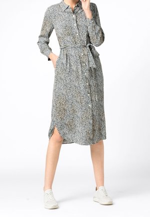 Femme portant une robe mi-longue à manches longues boutonnée, motif léopard, taille nouée, et des baskets blanches, debout sur fond blanc.