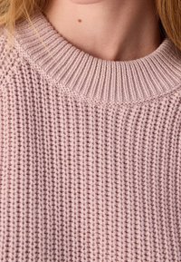 Gros plan sur une personne portant un pull tricoté rose pâle, montrant une texture détaillée et un grain de beauté visible sur la partie supérieure de la poitrine.