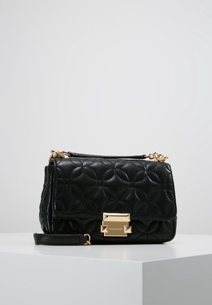 Černá prošívaná kožená kabelka se zlatou sponou a řetízkovým popruhem ležící na bílém povrchu na pozadí šedé stěny.