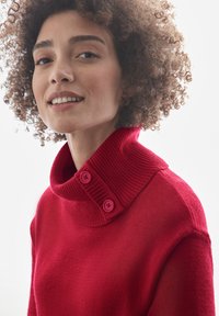 Roter Pullover mit hochgeripptem Kragen, der zwei pinke Knöpfe aufweist. Weiche Textur und taillierte Passform, die den Ausschnitt und die Schultern betont.