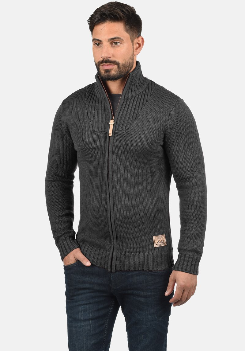 Cardigan lavorato a maglia di colore grigio scuro con collo alto, chiusura con cerniera, polsini a costine e un design testurizzato. Presenta una piccola patch con logo sull'orlo.