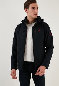 Melnā softshell jakā ar rāvējslēdzēju priekšpusē, kapuci un divām krūšu kabatām. Iezīmē sarkanas akcentus un elegantu, plūstošu dizainu.