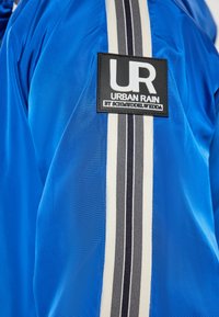 Giacca blu con finitura lucida, pannello laterale con strisce bianche e nere e un'etichetta con logo che recita "UR Urban Rain by Schmuddelwedda."