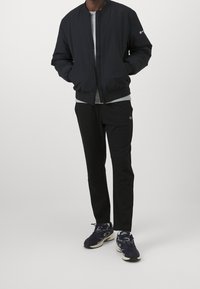 Person som bär en svart bomberjacka, grå skjorta, svarta byxor och svarta sneakers, står med händerna i jackfickorna.