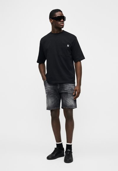 Črna majica s kratkimi rokavi z žepom na prsih, kombinirana z bledimi sivimi jeans kratkimi hlačami. Model nosi črna sončna očala in superge.