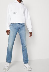 White hooded sweatshirt with "Calvin Klein Jeans" logo, paired with light blue straight-leg jeans and beige athletic shoes.