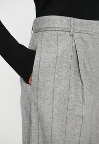 Personne portant un pantalon gris taille haute avec des coutures verticales et un haut noir à manches longues, main partiellement dans la poche du pantalon.