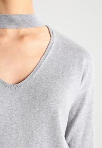 Lichtgrijze gebreide trui met een diepe V-hals en een vaste choker kraag, gedragen door een persoon tegen een effen achtergrond.