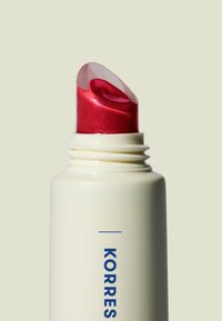 Produit à lèvres rouge scintillant dans un conteneur cylindrique blanc avec un capuchon transparent à vis. Le design présente des surfaces lisses et un texte graphique.