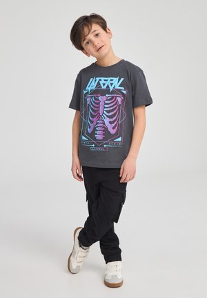 T-shirt grigia con grafica di uno scheletro colorato, pantaloni cargo neri e sneakers bianche con cinturini in velcro. Posizione in piedi contro uno sfondo semplice.
