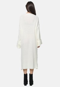 Lange witte gebreide cardigan met losse mouwen, gestructureerd patroon en een rechte snit, gedragen over zwarte enkelboots.