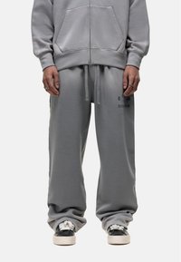 Light grey sweatpants with drawstring waist, loose fit, side pockets, and subtle branding on the left thigh. Soft cotton blend material.