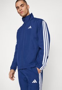 Μπλε αθλητικό μπουφάν με ψηλό γιακά, λευκές τρεις λωρίδες στα μανίκια, κλείσιμο με φερμουάρ και εμπρόσθιες τσέπες. Λογότυπο στο στήθος.