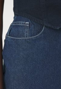 Tamsiai mėlynos džinsinės kelnės su tekstūruotu paviršiumi, turinčios galinį kišenę ir baltus siuvinėjimo akcentus. Viršuje matoma juosta.