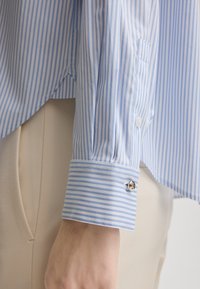 Blue and white striped shirt with buttoned cuff, featuring a smooth fabric texture and tailored design, accented by silver cufflink.