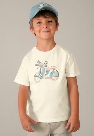 Ragazzo sorridente con un berretto azzurro chiaro e una maglietta bianca con una grafica di uno scooter pastello, in piedi davanti a uno sfondo semplice.