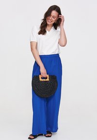 Weißes Kurzarmoberteil mit Kragen, kombiniert mit lockeren blauen Hosen. Das Model hält eine schwarze geflochtene Tasche mit einem Holzgriff.