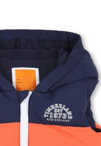 Giacca a cappuccio blu navy e arancione con fodera in pile, dotata di zip frontale, hardware bianco e logo del marchio sul petto.