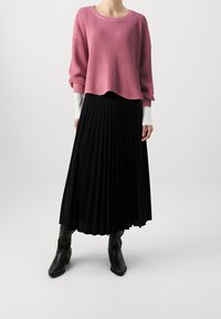 Rosa gerippter Pullover mit überschnittenen Schultern, kombiniert mit einem schwarzen plissierten Maxirock. Schwarze spitz zulaufende Stiefel vervollständigen das Outfit.