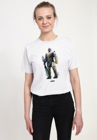 Biała bawełniana koszulka z ilustracją postaci w fioletowym i niebieskim zbroi, w czarnych dżinsach, na neutralnym tle.