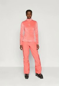 Nő korallszínű síkabátban és nadrágban, fekete síbakancsban áll egy világos szürke háttér előtt.