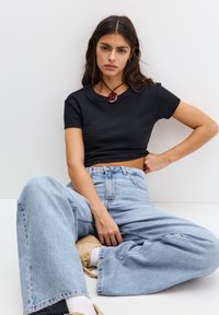 Zwarte geribbelde crop top, lichtblauwe wijde jeans, beige schoeisel, rode spiraal hangersnoer. Model zittend met een neutrale achtergrond.