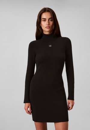 Frau mit langen dunklen Haaren, die ein eng anliegendes schwarzes, geripptes Langarmkleid mit hohem Kragen und kleinem Logo auf der Brust trägt.