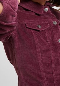 Burgundská kordová bunda s límečkem, dvěma předními kapsami a stříbrnými kovovými knoflíky. Textura je měkká s viditelným žebrováním.