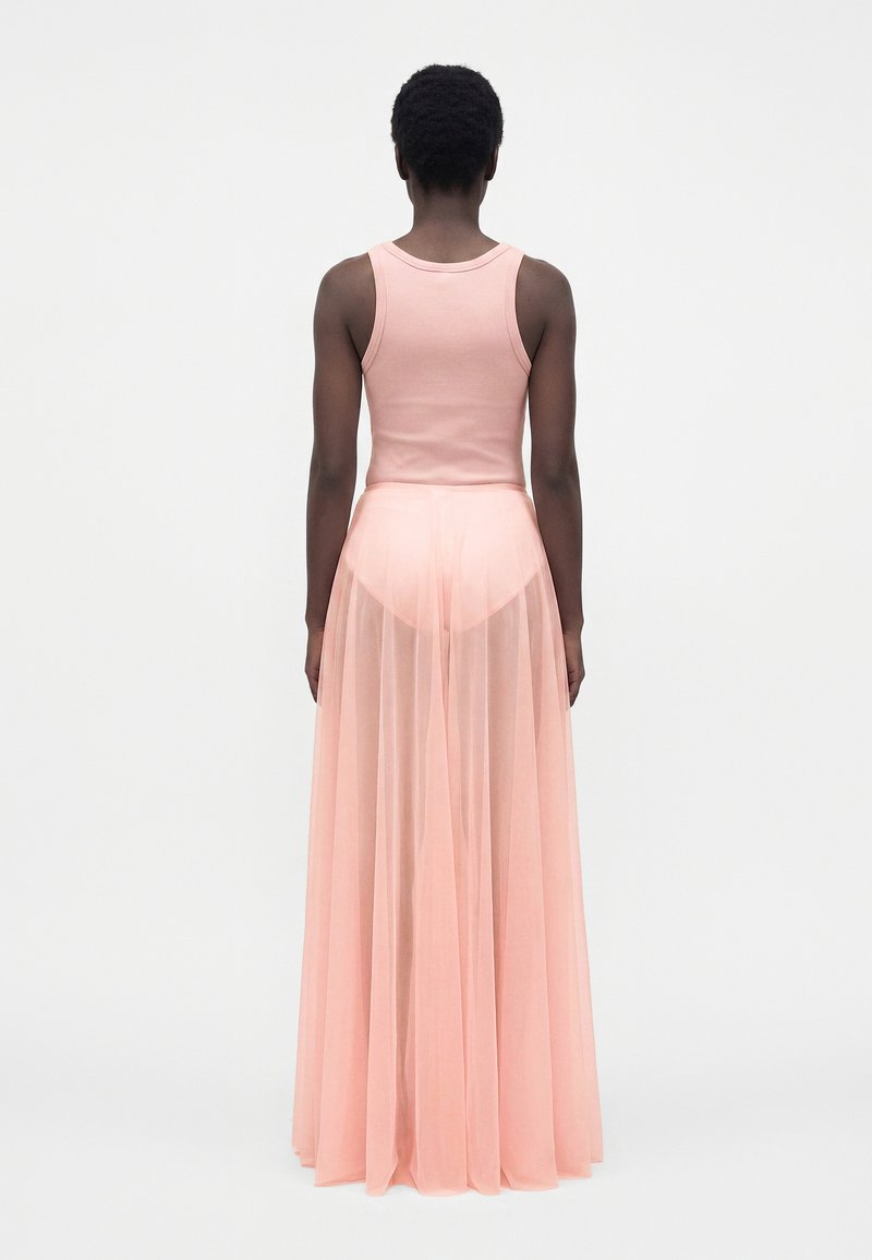 Personne aux cheveux courts et bouclés, vue de dos, portant un top rose sans manches et une longue jupe rose transparente par-dessus un short.