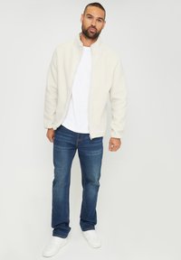 Giacca in fleece color crema con colletto alto e chiusura zip, abbinata a jeans blu e sneakers bianche, in piedi davanti a uno sfondo neutro.