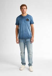 Camiseta azul degradada de mangas cortas, con escote redondo y detalle de parche circular, combinada con jeans de mezclilla claros y zapatillas blancas.