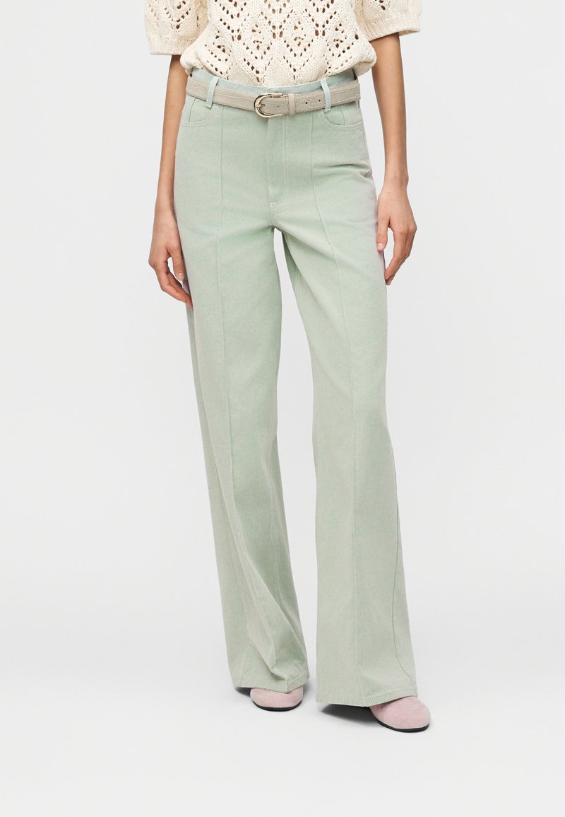 Γυναίκα που φοράει ανοιχτό πράσινο παντελόνι φαρδιάς γραμμής με μπεζ ζώνη, κρεμ πλεκτό τοπ και ανοιχτά ροζ παπούτσια, στέκεται μπροστά σε απλό φόντο.