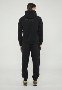 Sweatshirt à capuche noir avec logo EA7 dans le dos, accompagné de pantalons de jogging noirs assortis. Le tissu semble lisse ; le design est décontracté et ajusté.