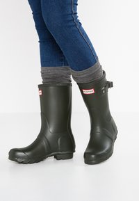Dunkelgrüne Gummiregenstiefel mit rundem Zehenbereich, strukturiertem Profil, verstellbarem Riemen und weißem Logoetikett. Getragen mit blauen Jeans und grauen Bündchen.