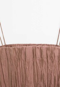 Tissu brun froissé avec fines bretelles spaghetti, encolure élastique froncée sur un vêtement sur fond blanc.