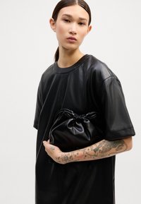 Camiseta oversized preta em couro com mangas curtas, combinada com uma pequena bolsa preta com cordão, apresentando uma textura suave e um design minimalista.