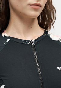 Gros plan d'une femme portant un haut noir zippé avec des accents floraux près des épaules et un petit logo blanc sur la poitrine.