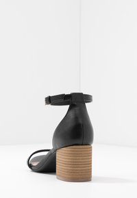 Sandale en cuir noir avec un bout arrondi, une bride de cheville réglable et un talon en bois cylindrique texturé.