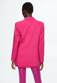Langer pinker Blazer mit strukturiertem Design und Reverskragen, der eine glatte Textur und lange Ärmel aufweist, getragen über engen pinken Hosen.