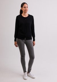 Cárdigan negro con botones, mangas largas, tela texturizada. Combinado con jeans gris ajustados y zapatillas blancas, de pie frente a un fondo liso.