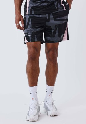 Atleet draagt zwarte en roze korte broek met patroon, witte kousen en wit-grijze hardloopschoenen, staande op een effen achtergrond.