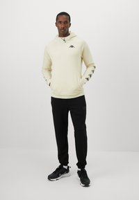 Man wearing beige hooded sweatshirt with logos on chest and sleeves, black joggers, and black sneakers, standing with hands in pockets.