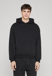 Giovane uomo con felpa nera semplice e pantaloni neri abbinati in piedi davanti a uno sfondo grigio chiaro.