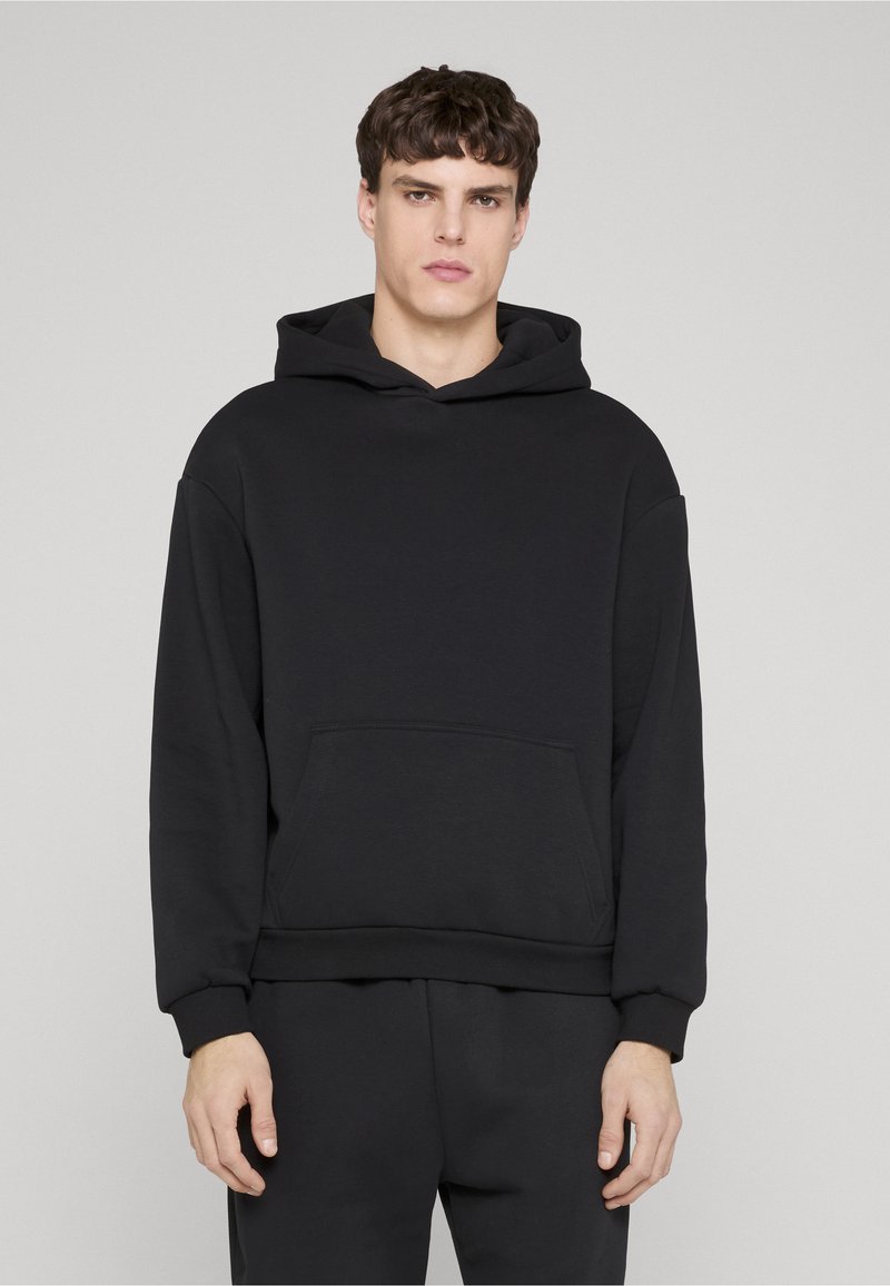 Giovane uomo con felpa nera semplice e pantaloni neri abbinati in piedi davanti a uno sfondo grigio chiaro.