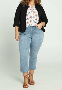 Blazer noir sur mesure, blouse blanche à imprimé coloré, jean clair coupé au-dessus de la cheville et talons bruns à brides.