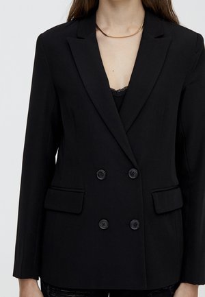 Woman wearing a black double-breasted blazer with buttons and flap pockets, paired with a black lace top and a gold and black chain necklace.