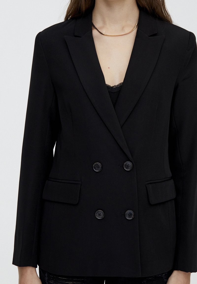 Femme portant un blazer noir croisé avec boutons et poches à rabat, associé à un top en dentelle noire et un collier chaîne or et noir.