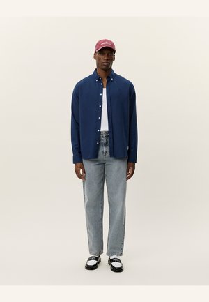 Camicia a bottoni blu navy, t-shirt bianca sotto, jeans azzurri chiari, scarpe in bianco e nero e un cappellino da baseball bordeaux. Completo semplice e casual.
