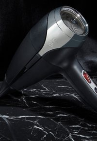 Sèche-cheveux ghd noir et argent avec bouton rouge, posé sur une surface en marbre noir sur fond sombre.